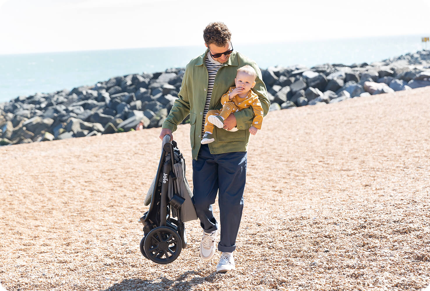 A dad walking on the beach carrying a baby on one arm and a folded litetrax 4 dlx stroller in the other hand.