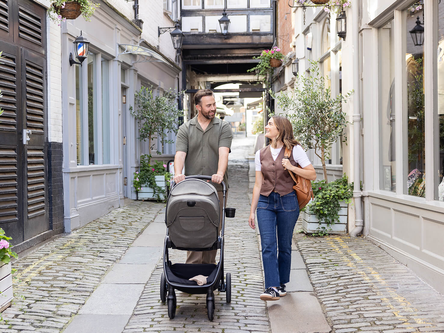 Parents pushing a Joie Signature finiti compact pram down a quaint cobblestone street filled with retail shops and greenery.
