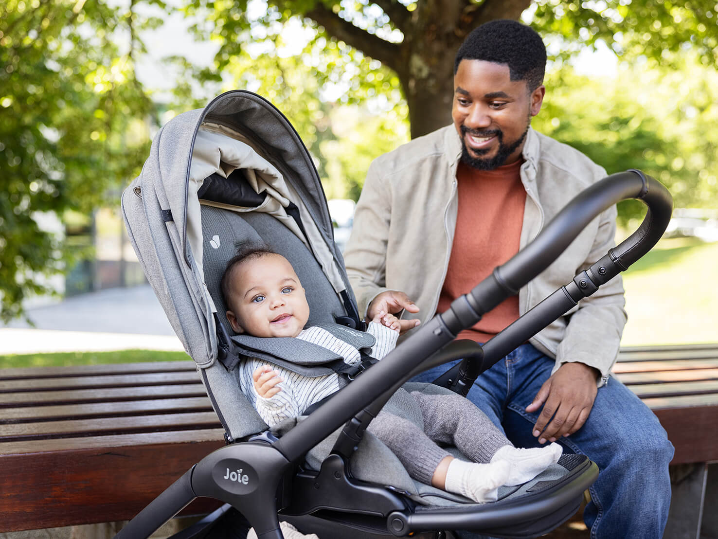 Dad playing with his baby in a Joie Signature finiti compact pram while sitting on a park bench.
