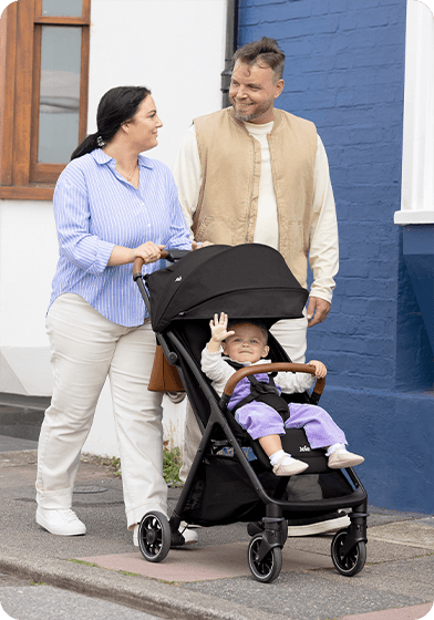 Mama und Papa gehen mit einem glücklichen Baby, das in einem schwarzen Joie pact pro sitzt, spazieren. 