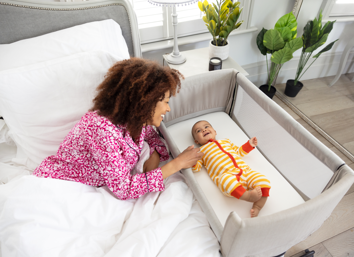 Mère penchée sur le côté de son lit et souriant à son bébé heureux, couché à plat dans la nacelle Joie Roomie Go, coloris gris. 