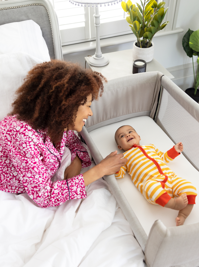 Mère penchée sur le côté de son lit et souriant à son bébé heureux, couché à plat dans la nacelle Joie Roomie Go, coloris gris. 