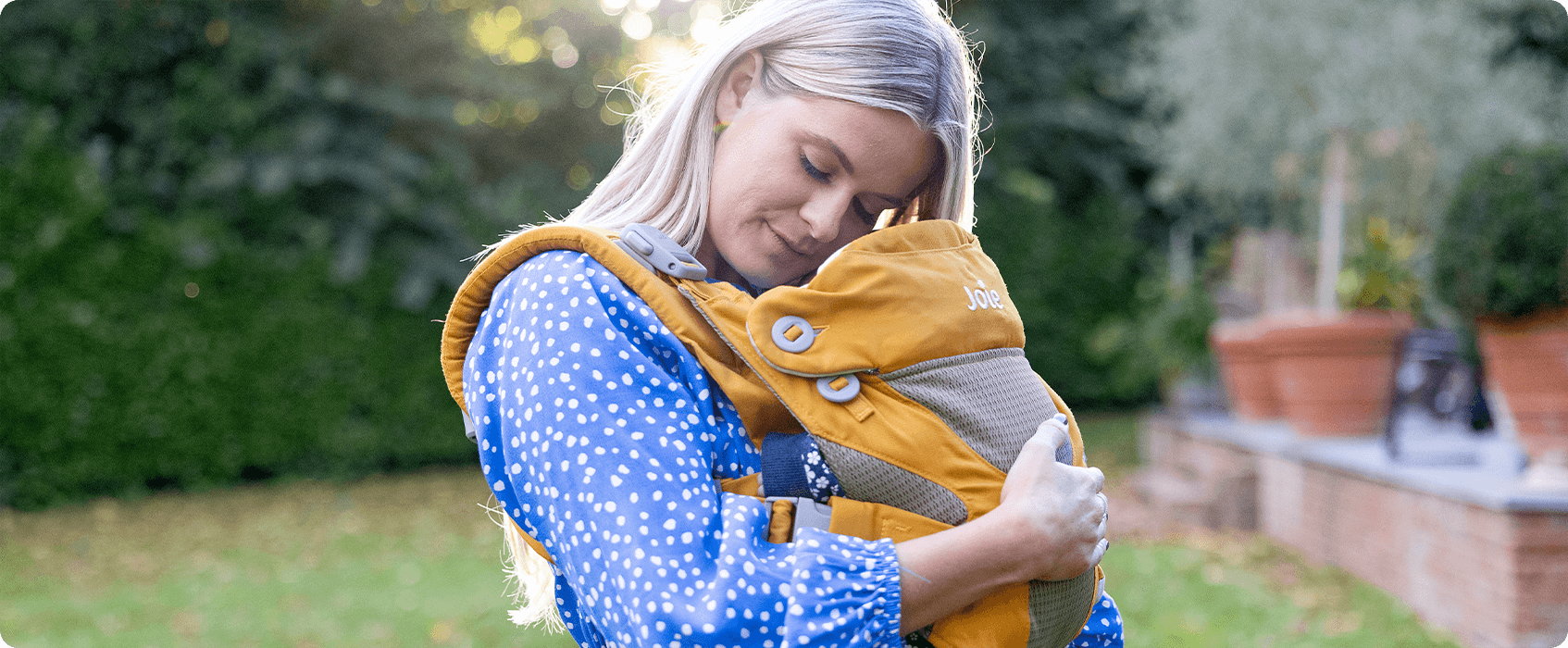 Mamá acurrucando a su bebé en un portabebés Joie Savvy amarillo mostaza